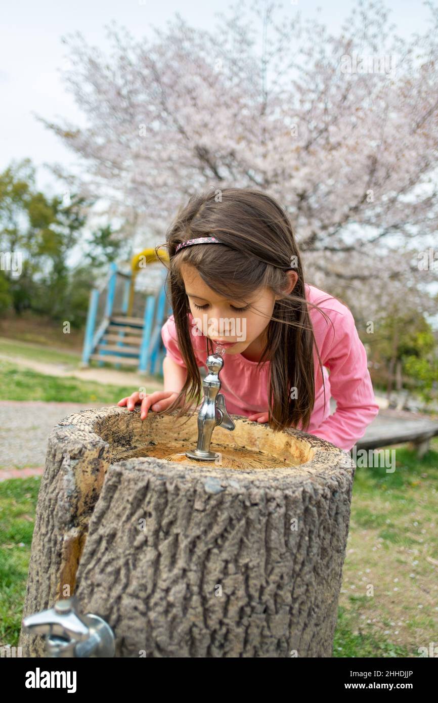 Bambina con lunghi capelli marroni che bevono acqua da una fontana nel parco. Primavera. Foto Stock