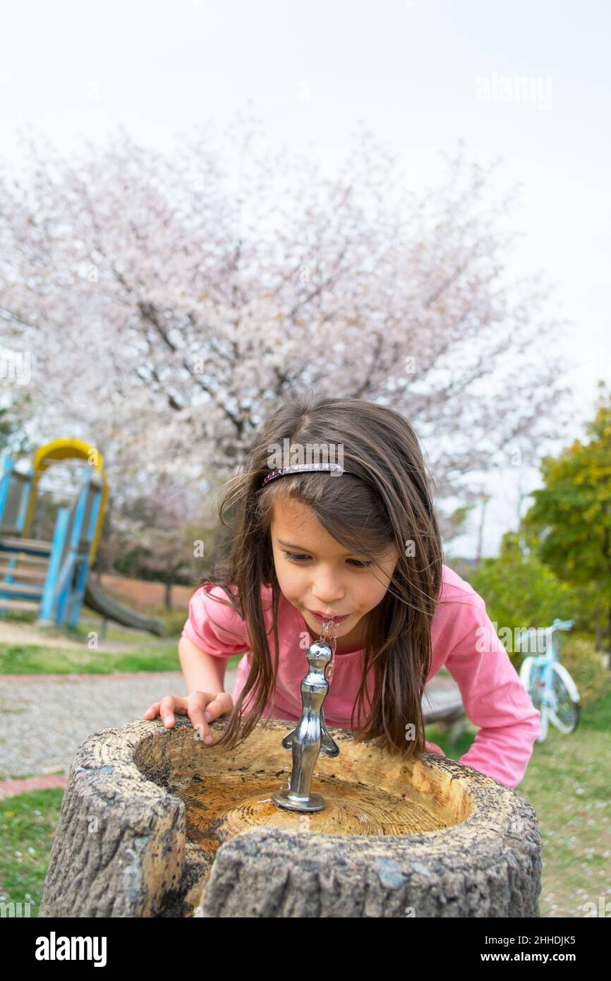 Bambina con lunghi capelli marroni che bevono acqua da una fontana nel parco. Primavera. Foto Stock