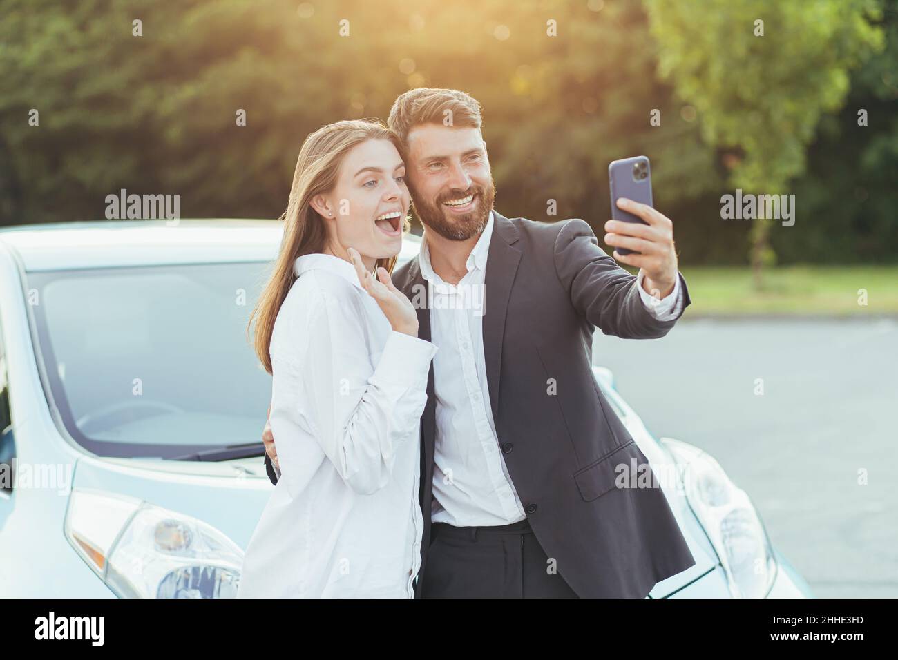 Giovane bella coppia uomo e donna, felice di acquistare una nuova auto, scattare foto insieme al telefono, e mostrare gli amici shopping tramite videochiamata Foto Stock