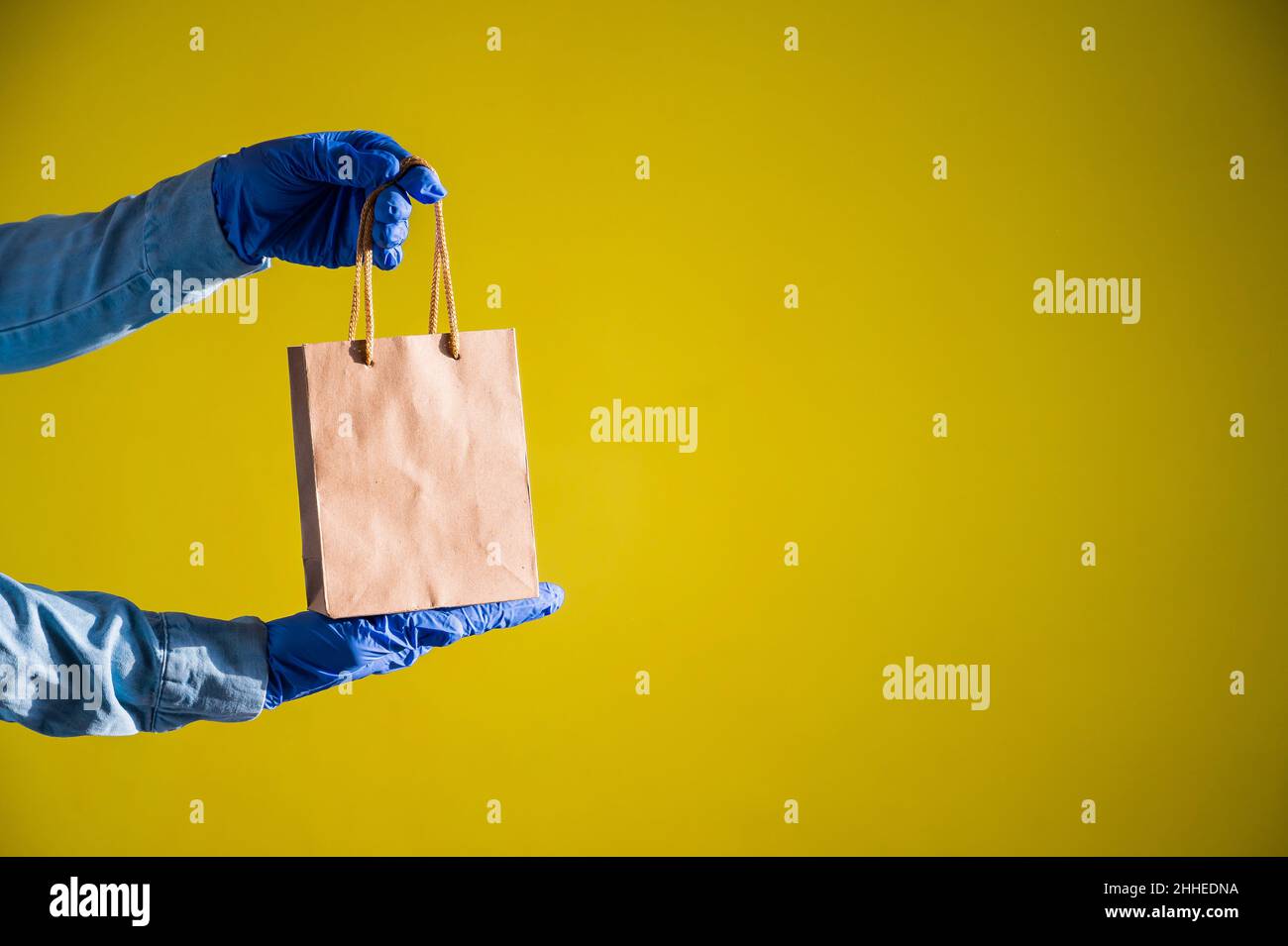 La mano del corriere femminile nei guanti medici del lattice contiene un sacchetto piccolo di carta. Somministrazione sicura durante la diffusione del coronavirus. Imballaggio ecologico. Online Foto Stock