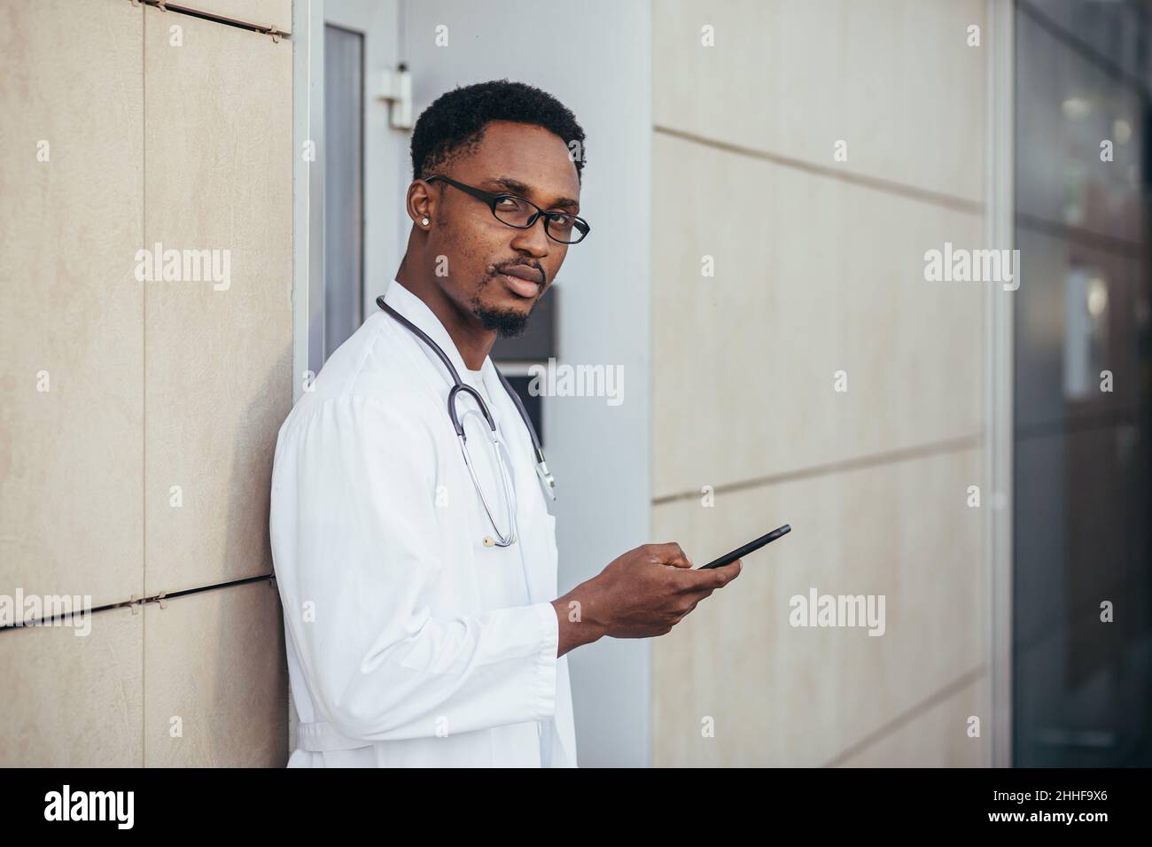 il medico afroamericano serio, vicino alla clinica, in un abito medico bianco guarda la macchina fotografica e tiene un telefono cellulare per parlare con i pazienti Foto Stock