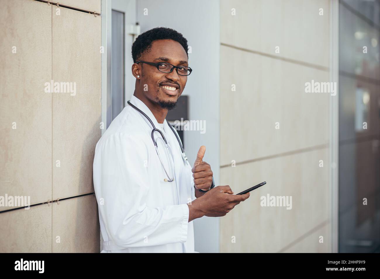Un uomo allegro di un medico afroamericano, vicino alla clinica, in un abito medico bianco guarda la macchina fotografica e tiene un telefono cellulare per parlare con il paziente Foto Stock