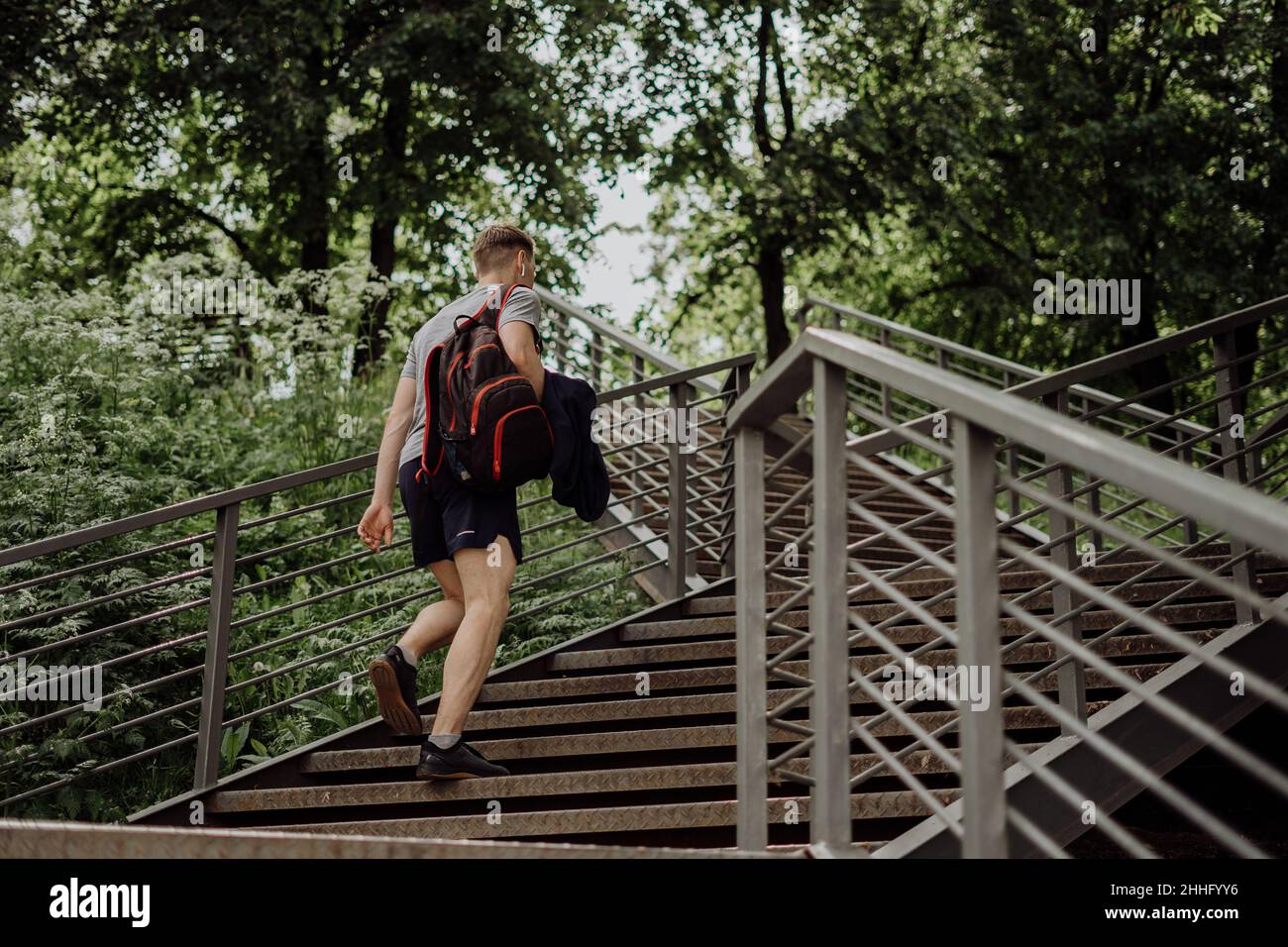 Stanco giovane runner in cuffie e zaino cammina e rilassante dopo l'allenamento sportivo. Tenere una bottiglia d'acqua mentre si fa esercizio in estate Foto Stock