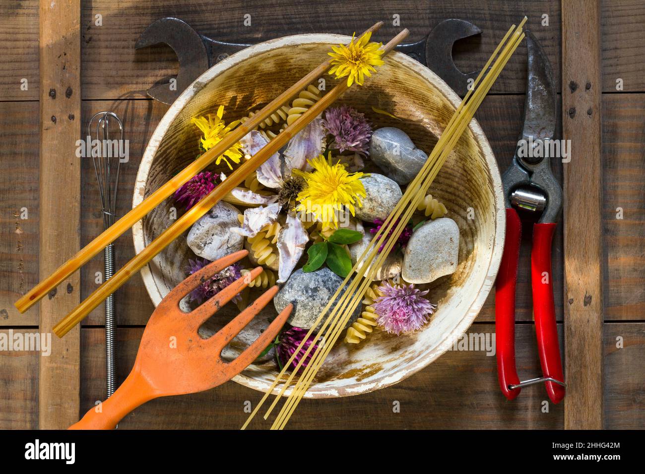 Piatto con pietre, fiori e utensili, bastoncini, spaghetti e pasta disposti come posate su un asse - immagine simbolica della vita morta di fame e povertà Foto Stock