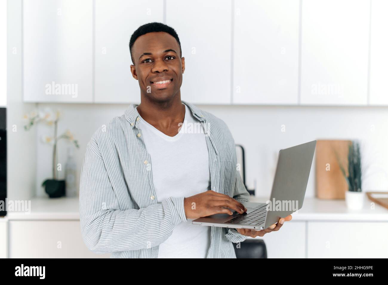 Bell'uomo, freelance o designer afro-americano giovane e sicuro, in piedi a casa in cucina, indossare abiti casual elegante, tenere un portatile aperto, guardare la macchina fotografica, sorridente amichevole Foto Stock