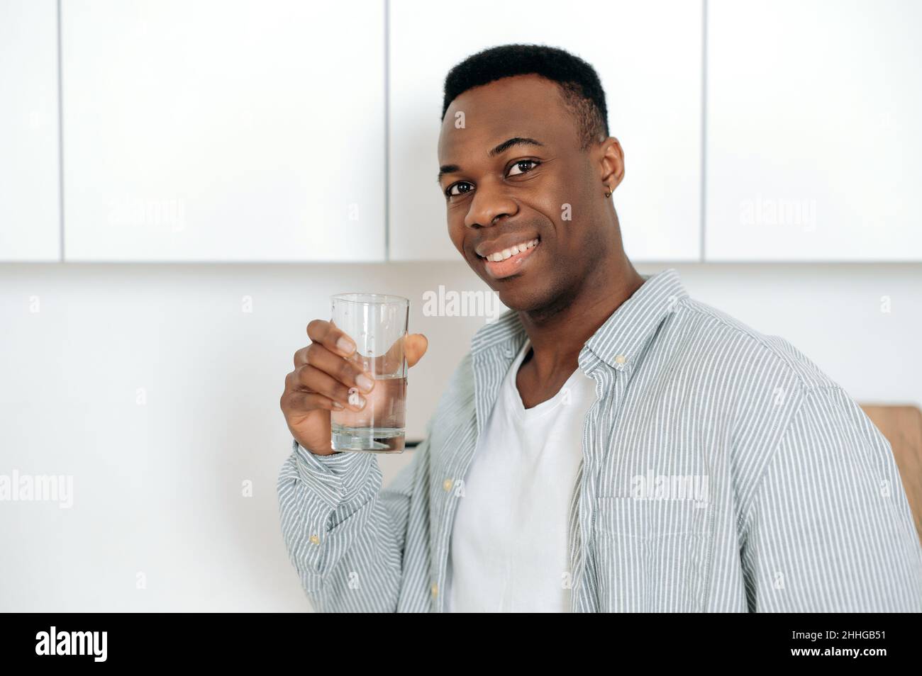 Concetto di assistenza sanitaria. Primo piano positivo afroamericano giovane uomo, in abbigliamento casual, si alza a casa in cucina, tiene un bicchiere di acqua pura, beve una quantità giornaliera di acqua, si prende cura di salute, sorriso Foto Stock