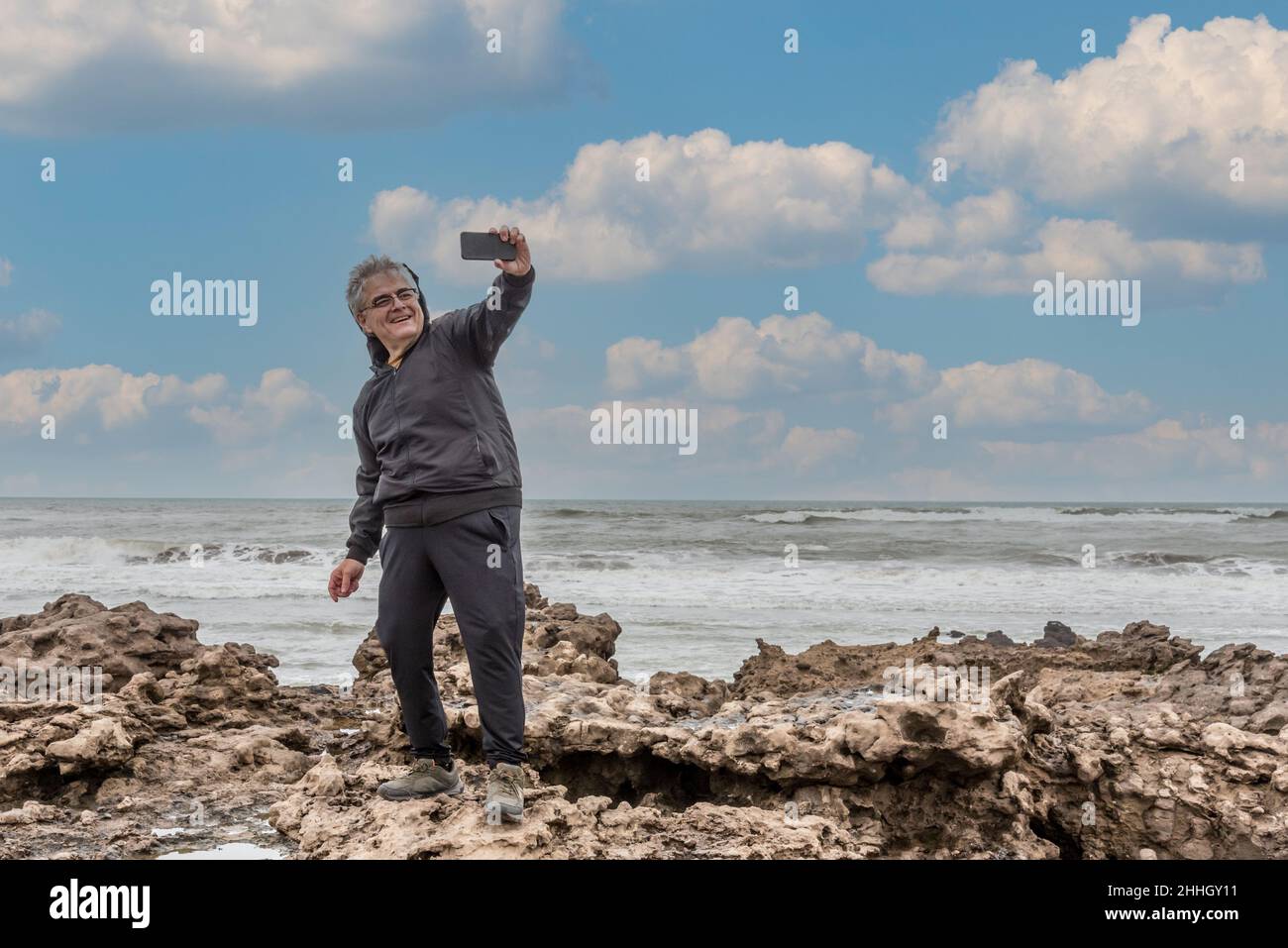 Turista adulto maturo con capelli e occhiali grigi prendendo un selfie sulle rocce con il mare alle spalle e un cielo con le nuvole. Foto Stock