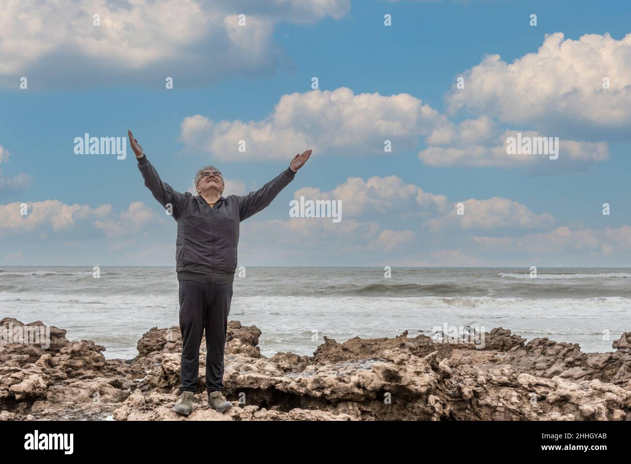 Uomo adulto con capelli e occhiali grigi che alzano le braccia al cielo sopra le rocce e il mare dietro. Foto Stock