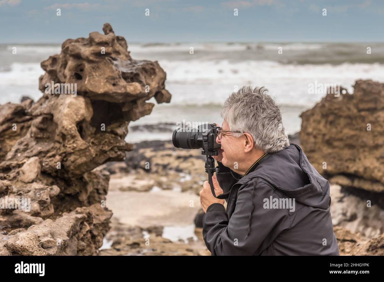 Uomo adulto maturo dai capelli grigi con occhiali che scattano una foto vicino alla costa marina. Foto Stock