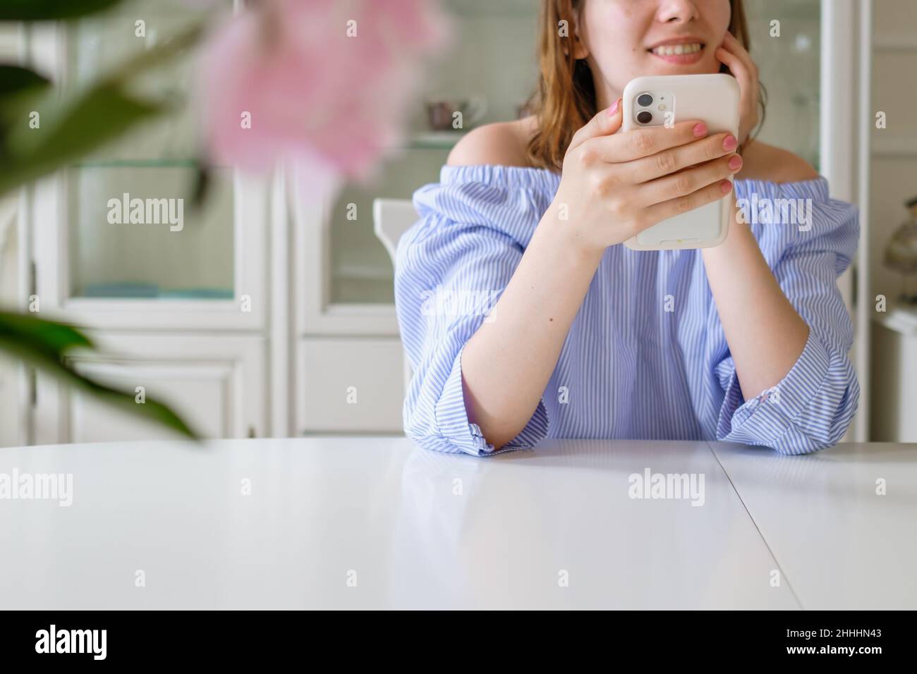 Allegra donna elegante in blusa blu controlla i social network in smartphone seduto al tavolo bianco con fiori in primo piano a casa primo piano Foto Stock