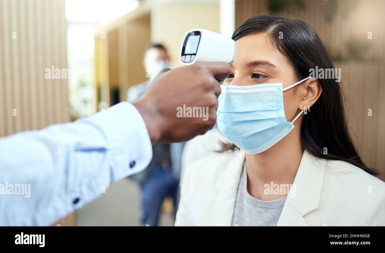 Prendiamo la temperatura. Scatto corto di una giovane donna d'affari attraente che indossa una maschera e che ha la sua temperatura presa mentre si levano in piedi al Foto Stock