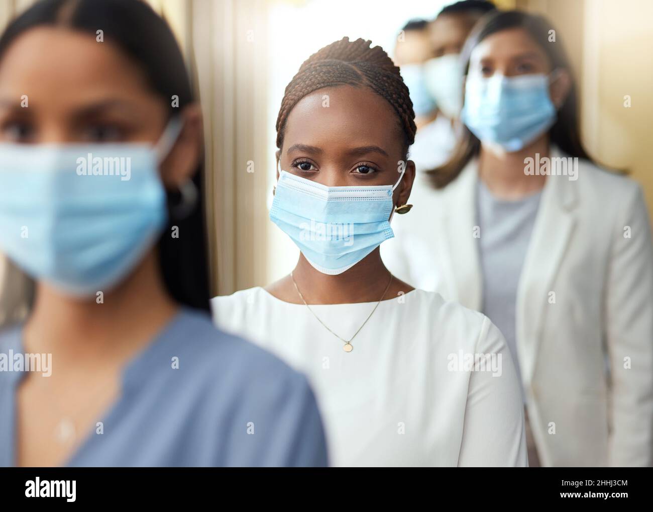 Accesso sicuro al luogo di lavoro. Scatto corto di una giovane donna d'affari attraente che indossa una maschera mentre sta in piedi nel mezzo di una coda nel suo ufficio. Foto Stock