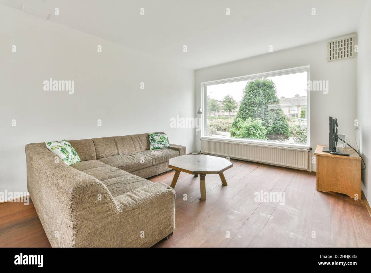 Vista del soggiorno bianco in stile minimale con televisore, divano, tavolo in ottagono di legno e una grande finestra Foto Stock