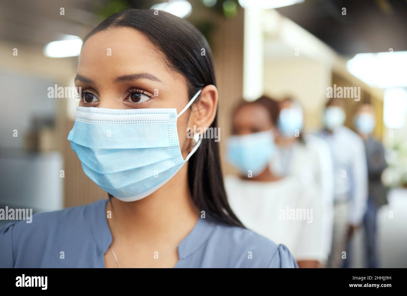 Attenzione pratica. Scatto corto di una giovane donna d'affari attraente che indossa una maschera mentre si trova in piedi alla testa di una coda nel suo ufficio. Foto Stock