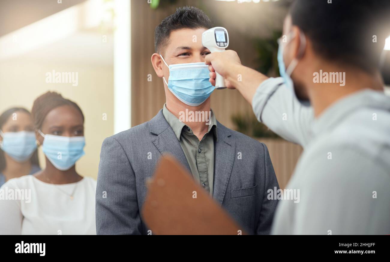 Allineati per essere schermati. Scatto corto di un bel uomo d'affari maturo che indossa una maschera e che ha la sua temperatura presa mentre sta in piedi alla testa di un Foto Stock