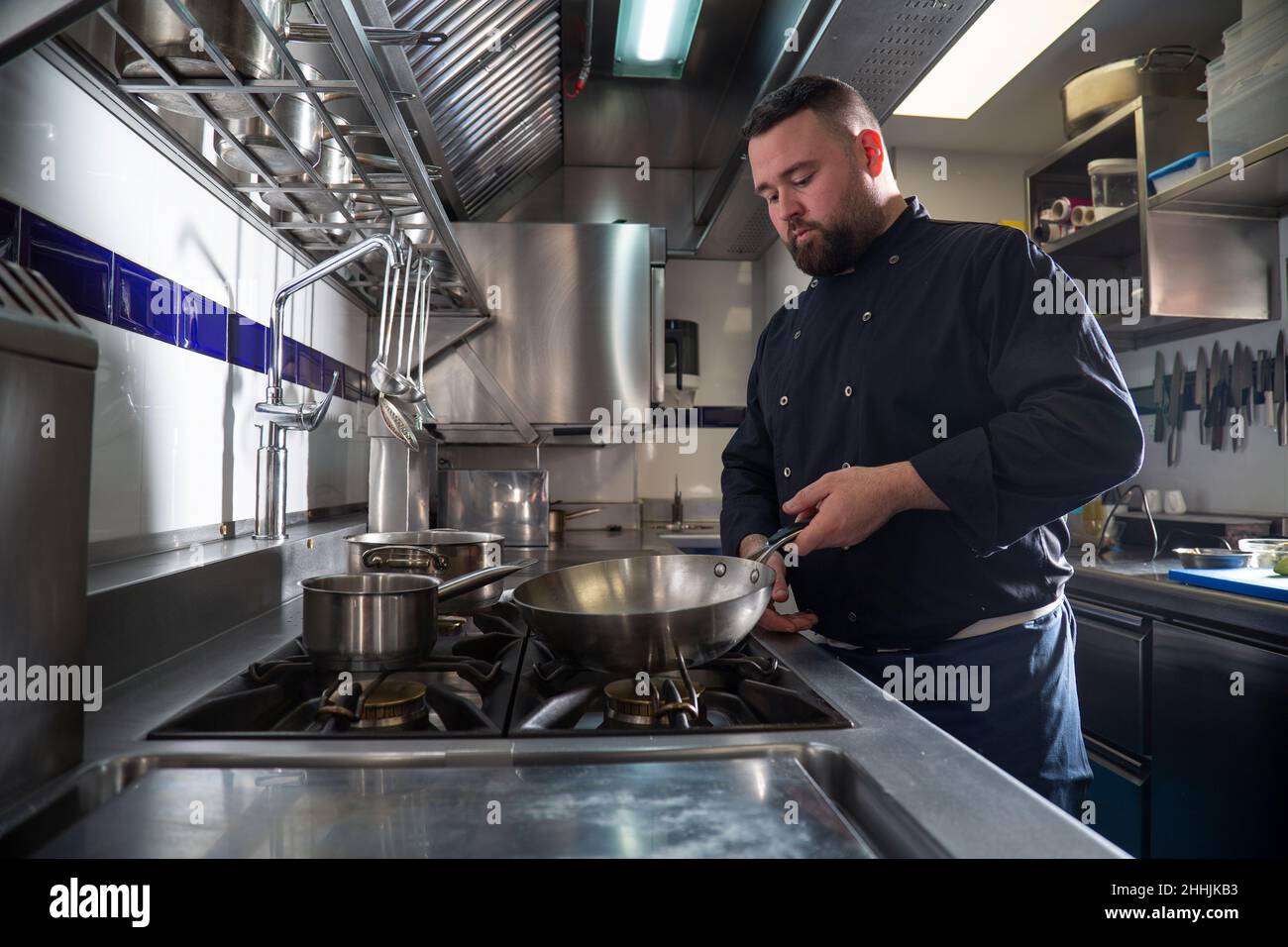 L'uomo in grumo in uniforme scuro mette la padella sul piano di cottura mentre cucinano il piatto nella cucina contemporanea del ristorante Foto Stock