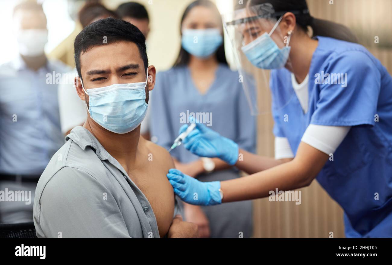 Non gli piacciono gli aghi. Scatto corto di un bel giovane uomo d'affari che ottiene la sua vaccinazione covid da un'infermiera femminile in ufficio. Foto Stock