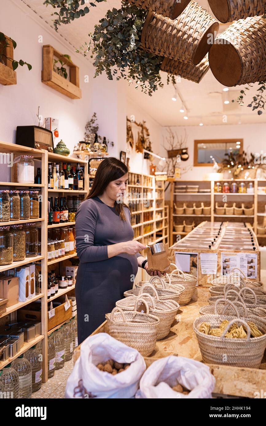 Vista laterale della donna incinta con capelli marroni in abito viola in piedi e la scelta dei prodotti nel negozio ecologico Foto Stock