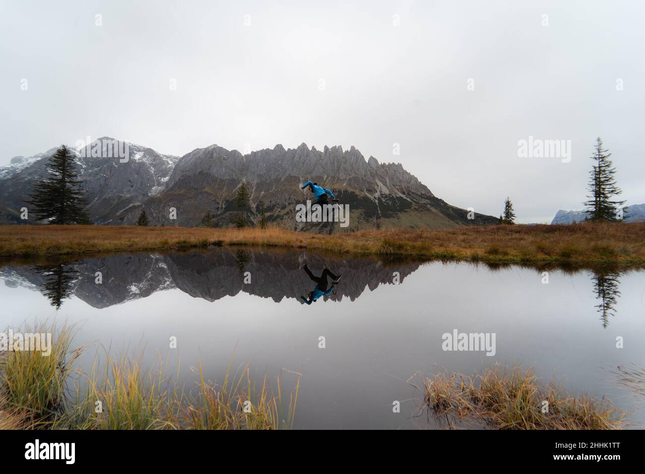 Lontano viaggiatore maschio che salta sulla riva erbosa del lago calmo vicino cresta di montagna in una giornata di nebbia in Austria Foto Stock