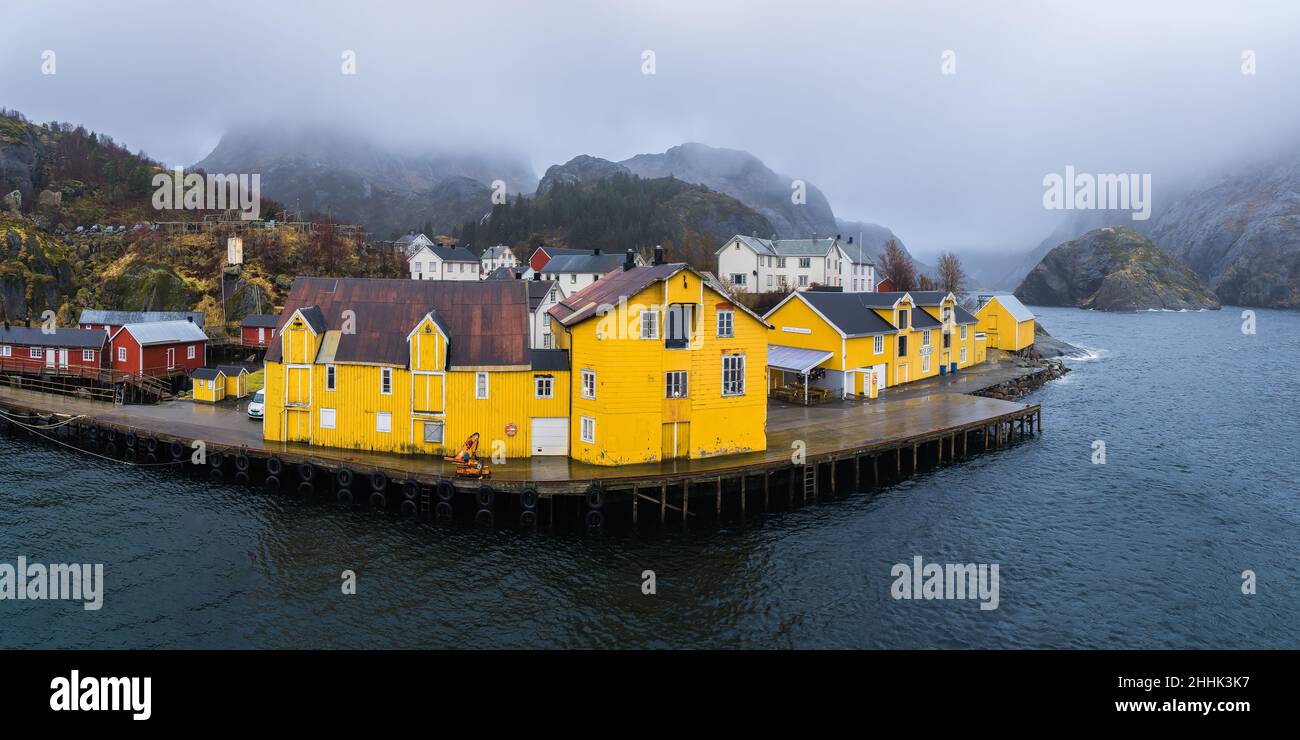 Vista panoramica del drone di un piccolo insediamento con tipiche case residenziali colorate al mare circondato da enormi montagne rocciose sotto il cielo nebbia a Nusfjo Foto Stock