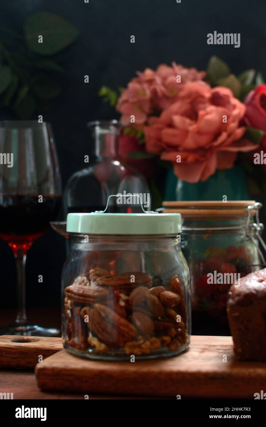 Vaso trasparente di vetro riempito con noci posto su tagliere di legno sul tavolo con alcol e fiori in fiore in cucina Foto Stock