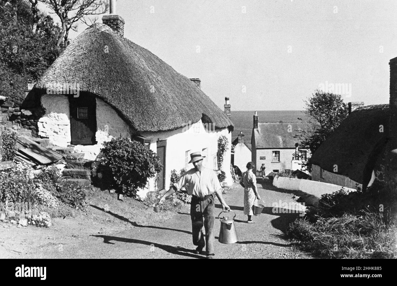 Un uomo trasporta un churn di latte su il farafare principale in Clovelly. Il villaggio è adagiato su una ripida collina e ha solo una strada acciottolata singola alta che si snoda lungo la collina attraverso tradizionali cottage imbiancati di bianco festoonati con fuchsias e gerani. Circa agosto 1935 Foto Stock