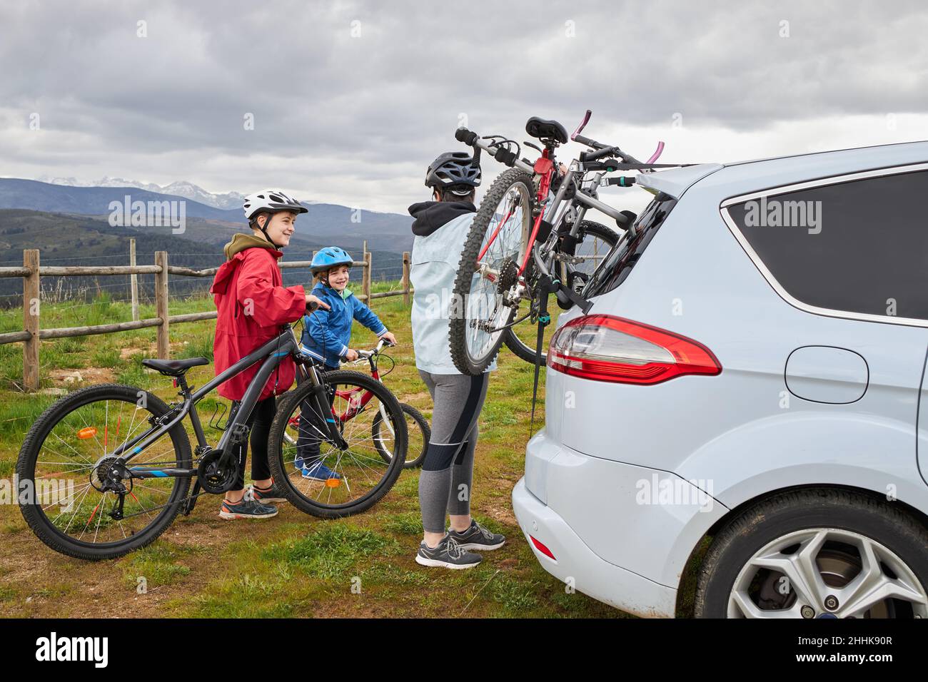 Vista laterale della madre positiva con i bambini in caschi che prendono la bicicletta dal portabagagli della macchina moderna parcheggiata in campagna Foto Stock