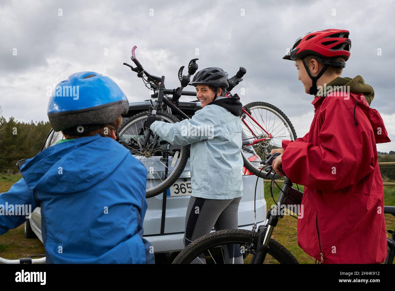 Madre positiva con i bambini in caschi che prendono la bicicletta dal portabagagli della macchina moderna parcheggiata in campagna Foto Stock
