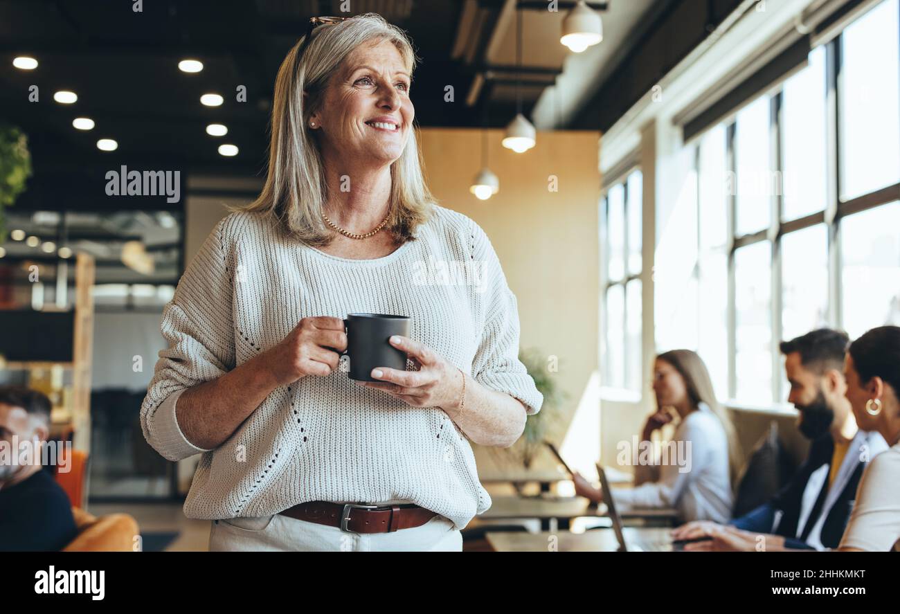 Donna d'affari pensata in piedi in uno spazio di co-lavoro. Donna d'affari matura sorridendo allegro mentre tiene una tazza di caffè. Entreprene esperto Foto Stock