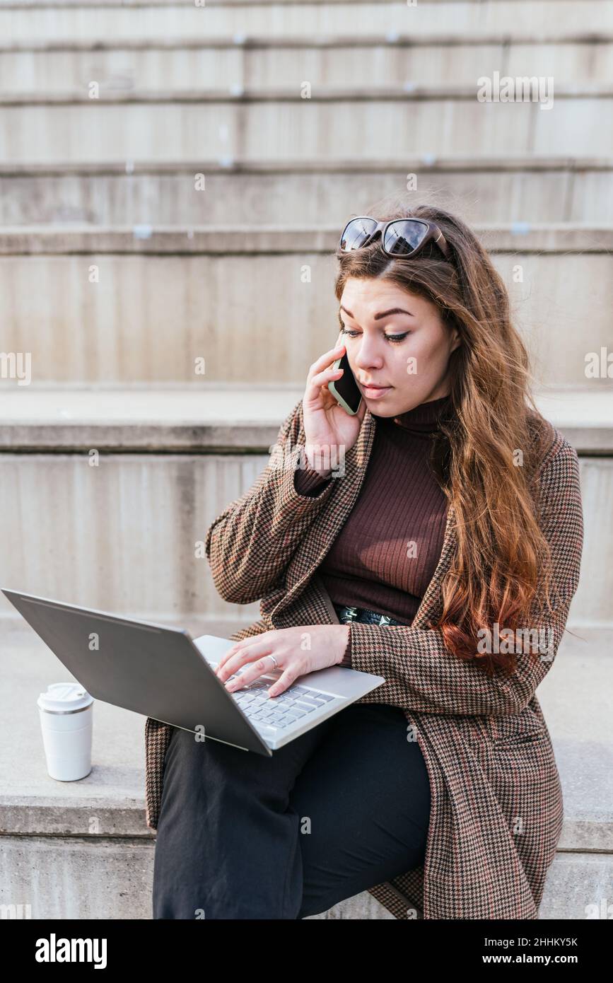 Donna freelancer in outerwear con capelli lunghi. Dati di navigazione su netbook e parlare sul cellulare mentre si siede sulle scale vicino al caffè per andare in città strada Foto Stock