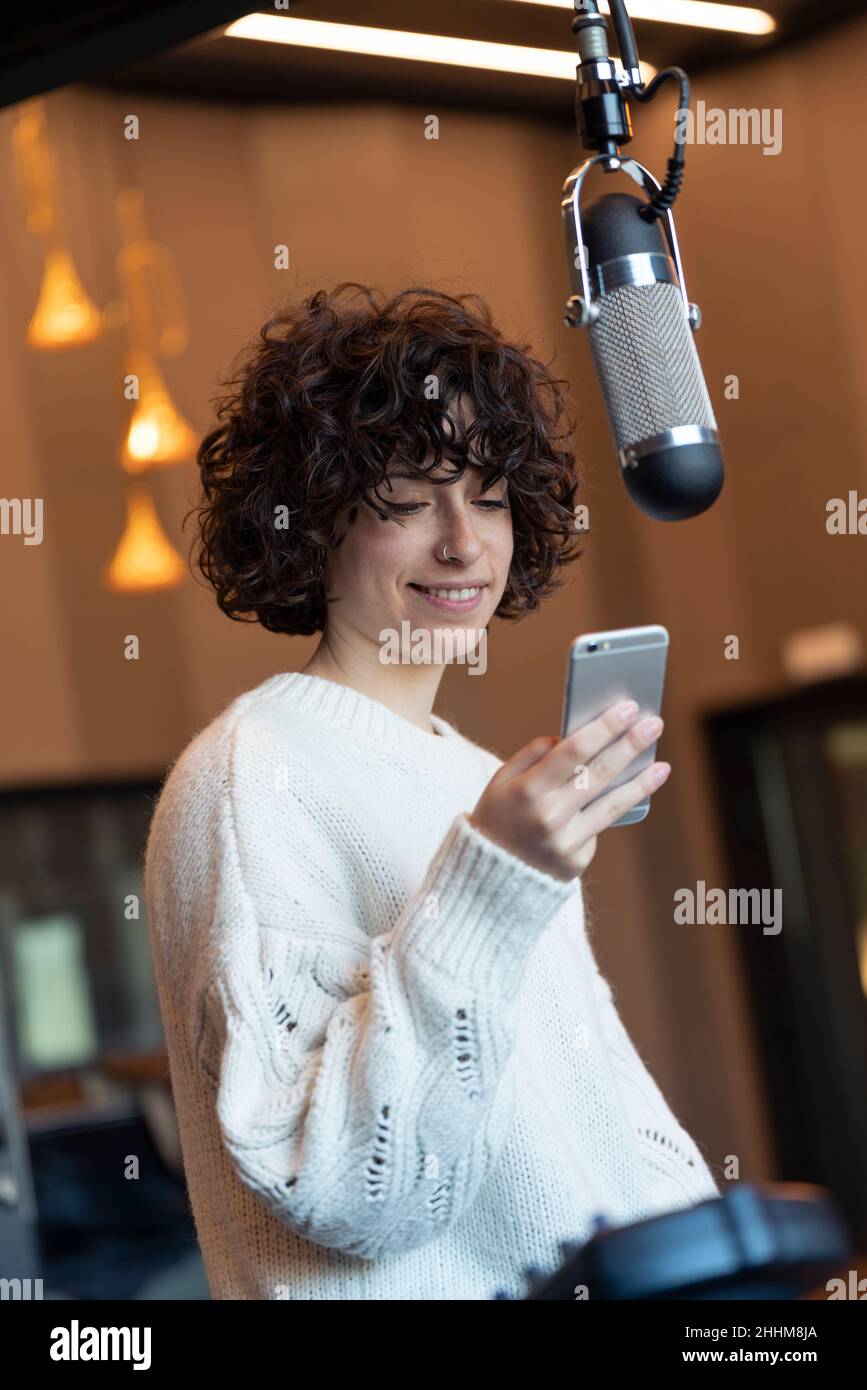 Caucasica giovane capelli ricci cantante donna sorridente con il suo telefono in uno studio di musica Foto Stock