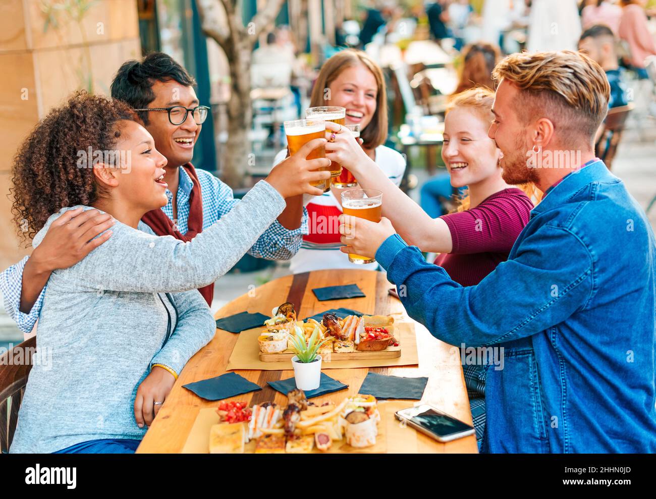 Amici multirazziali soddisfatti seduti al tavolo e guardandosi l'un l'altro mentre si aggraffano bicchieri con birra nella caffetteria di strada Foto Stock