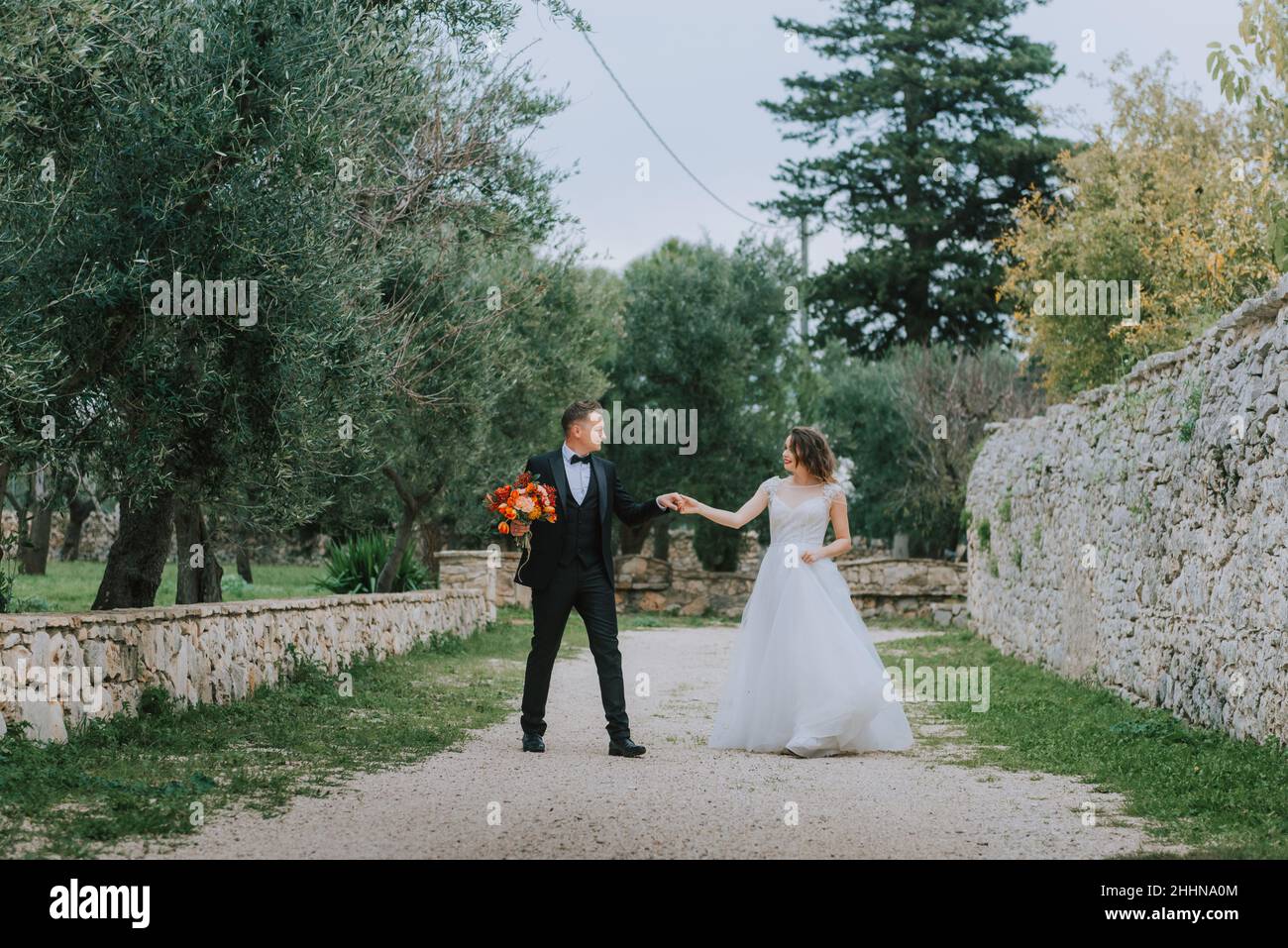 Felice coppia sorridente di stile che cammina in Toscana, Italia il giorno del matrimonio. La sposa e lo sposo camminano lungo la strada per le mani. Un giovane di stile Foto Stock