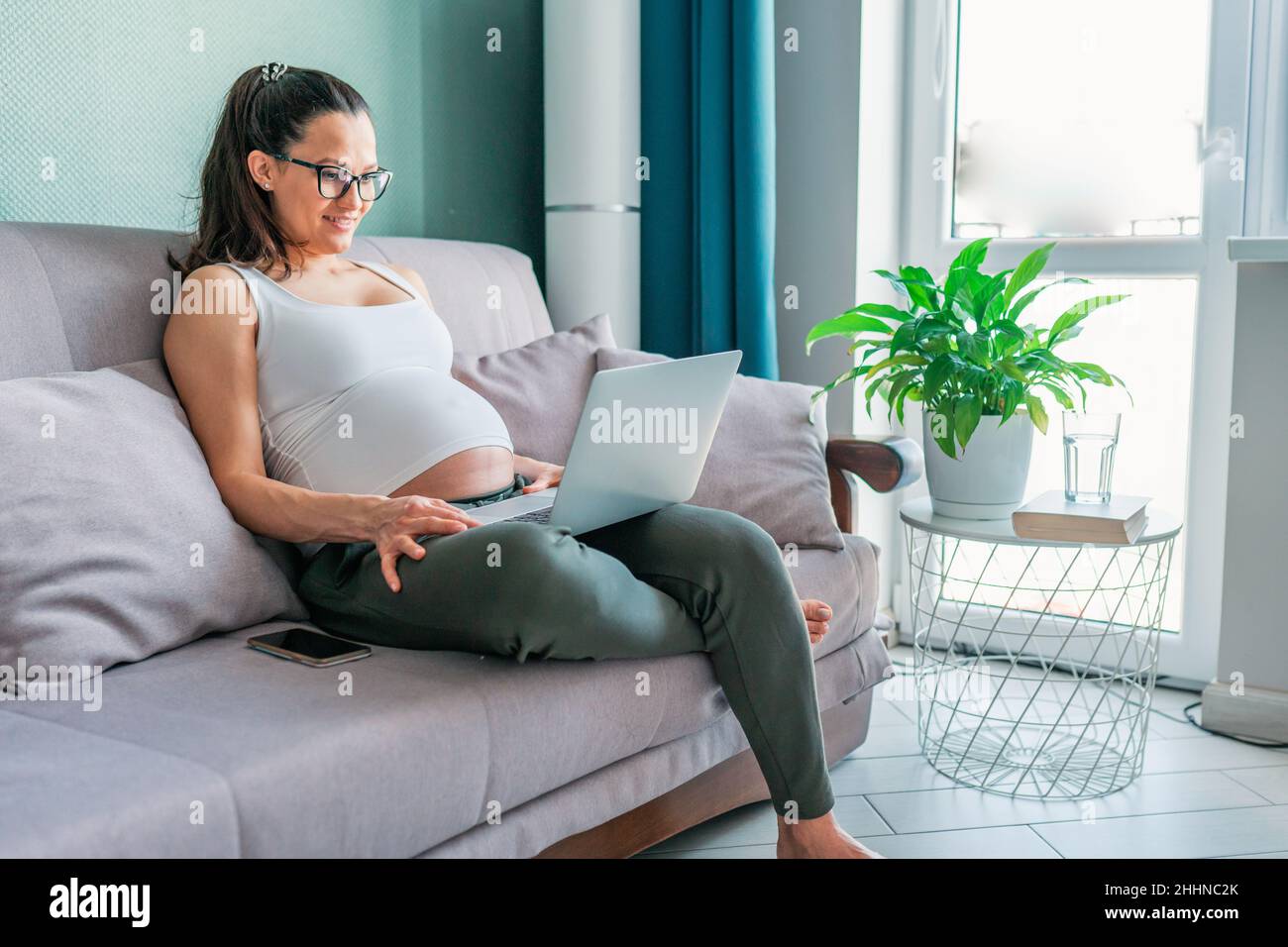 Donna d'affari incinta con computer portatile in salotto lavoro a casa distanza sul divano Foto Stock