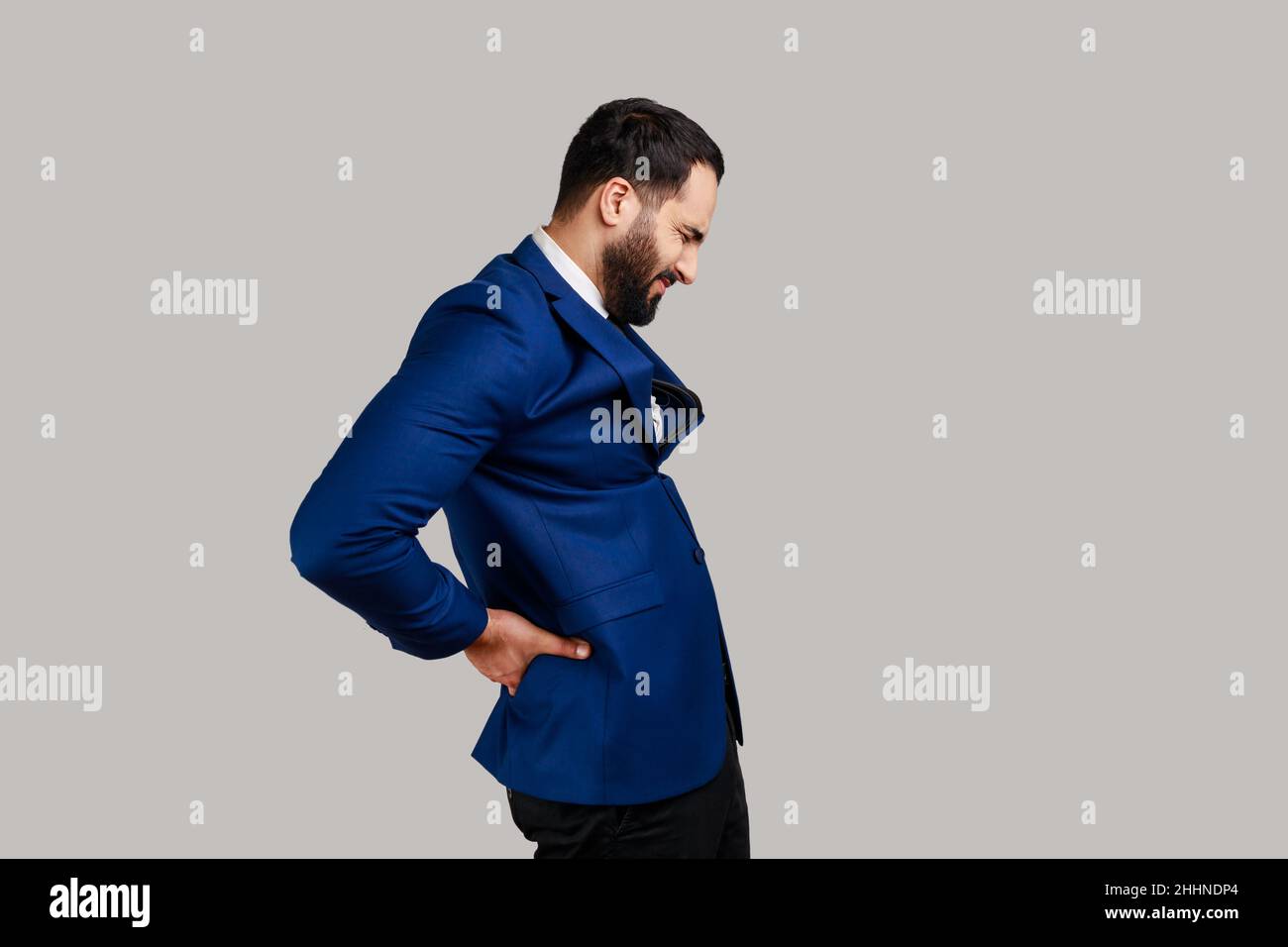 Ritratto della vista laterale dell'uomo barbuto in piedi con gli occhi chiusi, che soffre di dolore alla schiena più basso, infiammazione renale, vestito di stile ufficiale. Studio interno girato isolato su sfondo grigio. Foto Stock