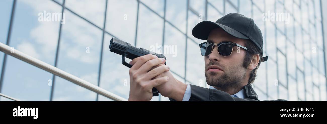 giovane uomo di sicurezza in berretto nero e occhiali da sole che tengono la pistola all'aperto, banner Foto Stock