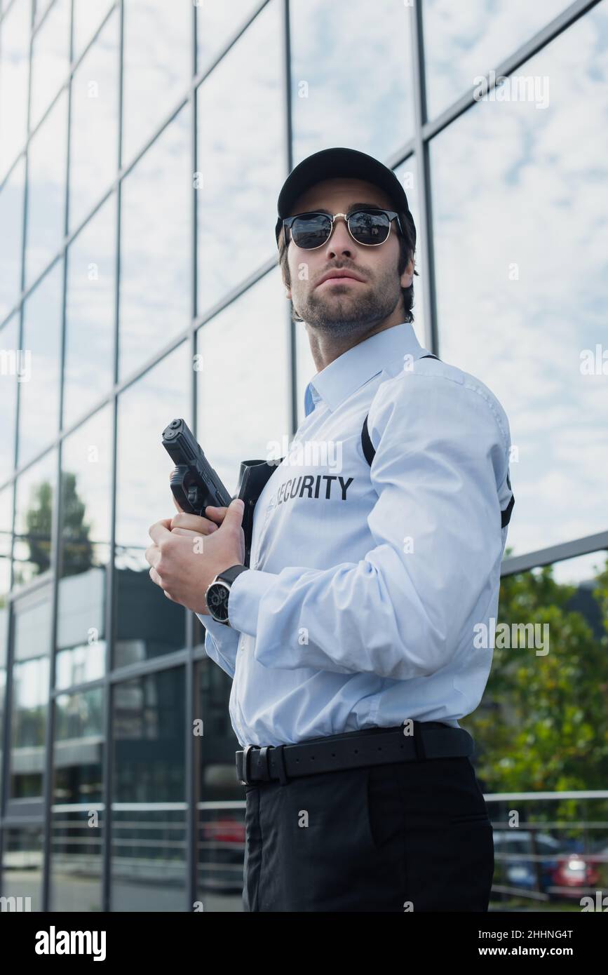 giovane guardia in occhiali da sole e cappuccio nero in piedi con pistola vicino edificio con facciata di vetro Foto Stock