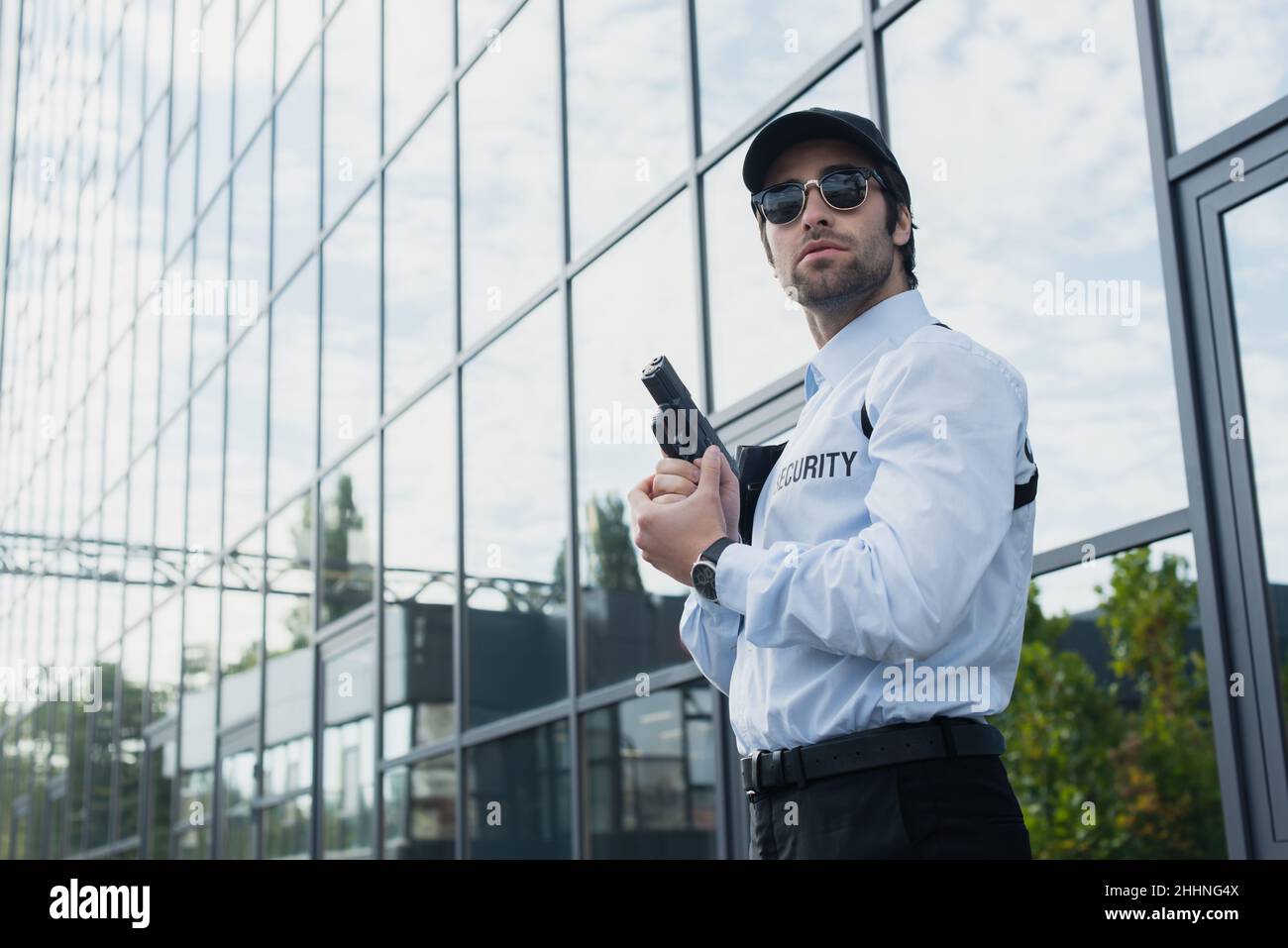 uomo di sicurezza in occhiali da sole in piedi con arma vicino edificio con facciata di vetro Foto Stock