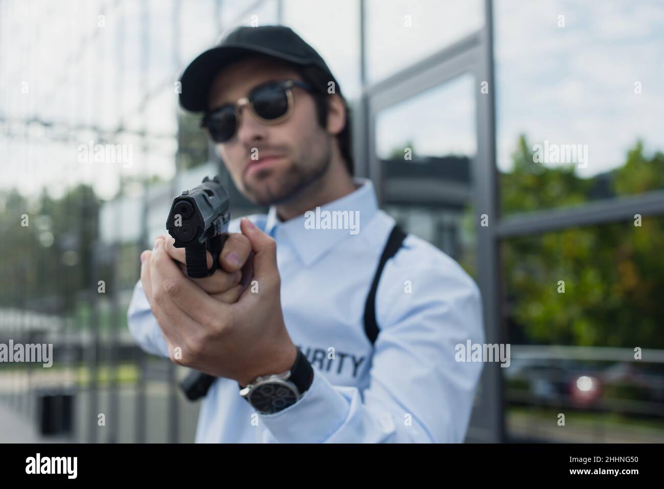 uomo di sicurezza offuscato in cappello e occhiali da sole che reggono la pistola all'aperto Foto Stock