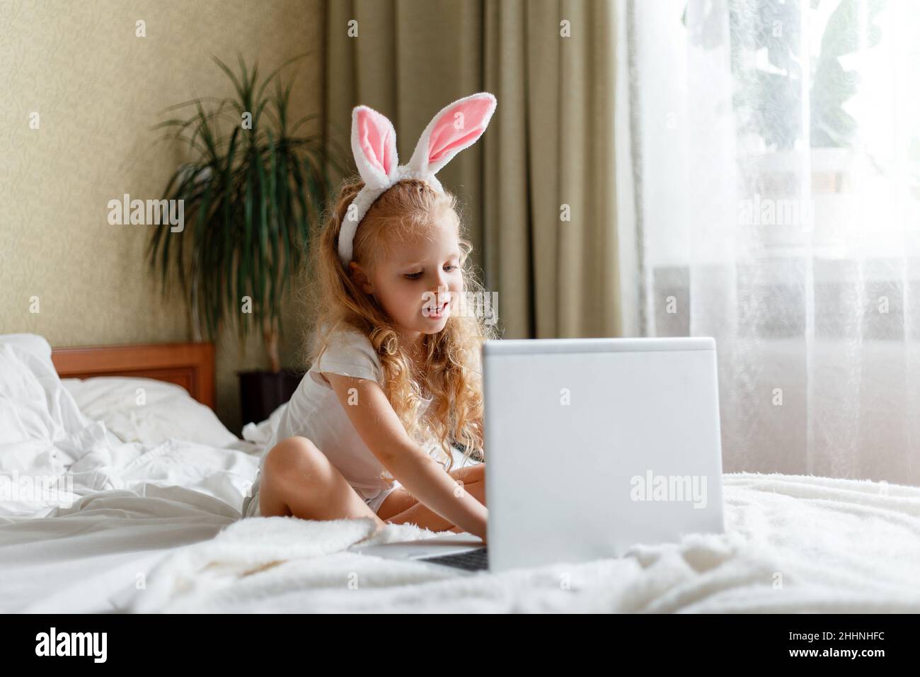 La ragazza divertente piccola felice usa il laptop per le videochiamate. Caucasico biondo curly bambino in coniglietto orecchie studiare a distanza giocare o guardare video via laptop in Foto Stock