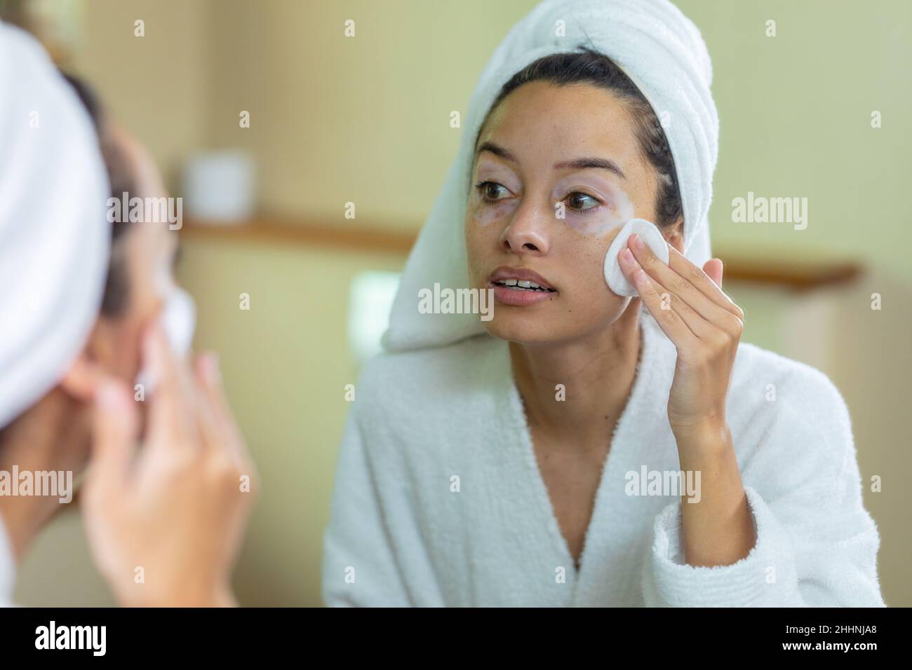 Giovane donna biraciale che indossa un accappatoio che applica crema sul viso davanti allo specchio in bagno. Inalterato, di routine, stile di vita e vitiligo concetto. Foto Stock