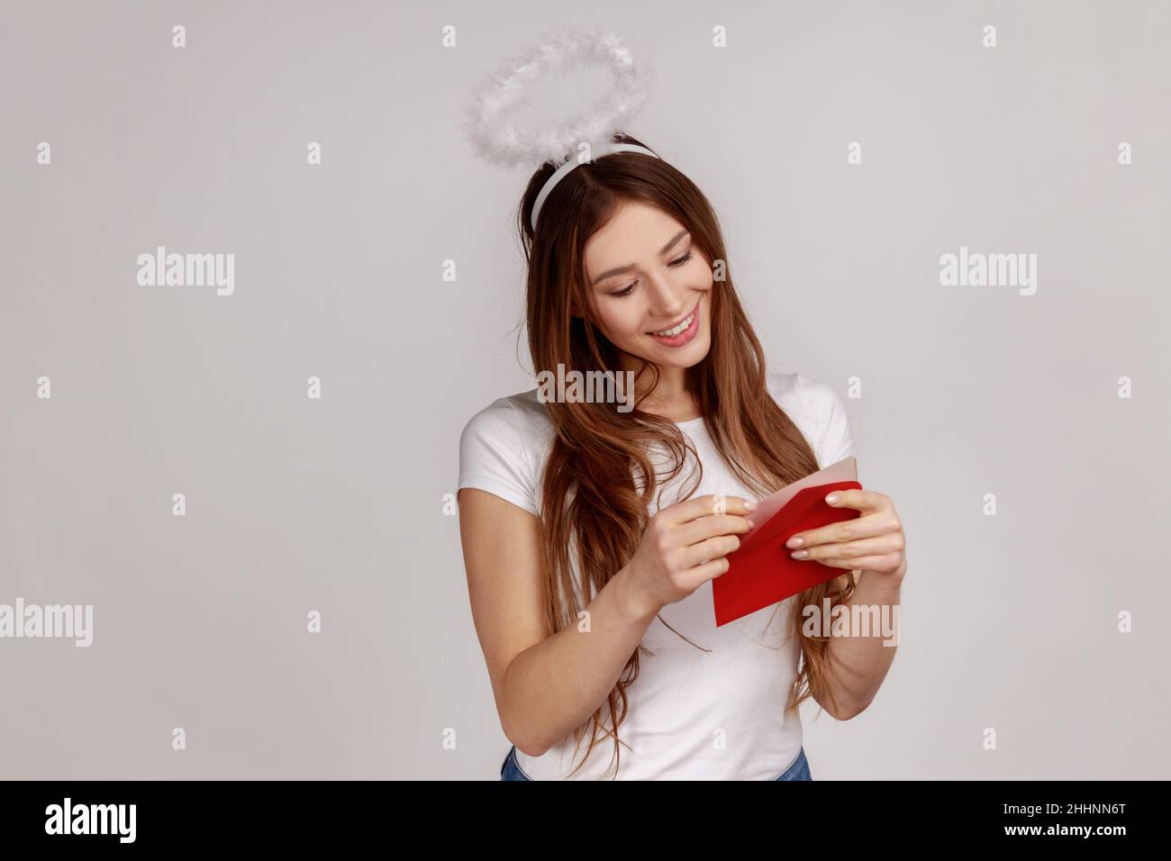 Ritratto di affascinante donna compiacente con nimbus sopra la testa che tiene busta rossa, lettura lettera romantica con sorriso, indossando T-shirt bianca. Studio interno girato isolato su sfondo grigio. Foto Stock