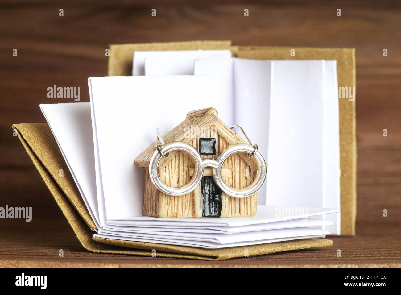 Modello di casa in miniatura che indossa occhiali in acciaio in piedi su un libro aperto con pagine bianche. Concetto creativo di contabilità domestica. Foto Stock