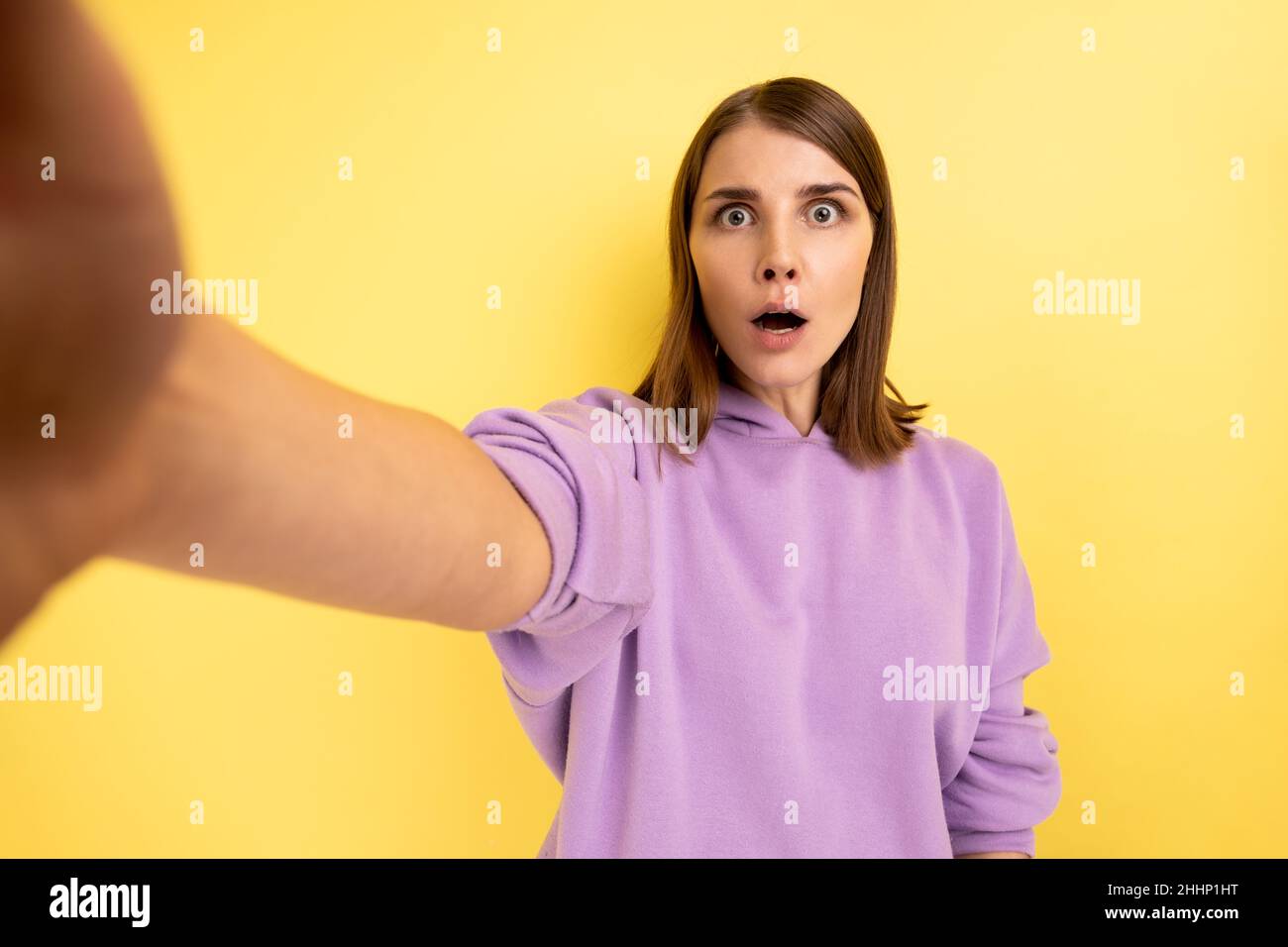 Ritratto di donna sorpresa prendendo selfie con espressione facciale scioccata, guardando con occhi grandi e bocca aperta, POV, indossando felpa con cappuccio viola. Studio interno girato isolato su sfondo giallo. Foto Stock