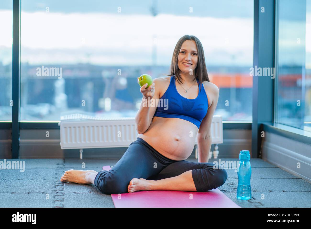 Donna incinta felice seduta sul tappeto yoga con mela mentre si esercita in palestra Foto Stock