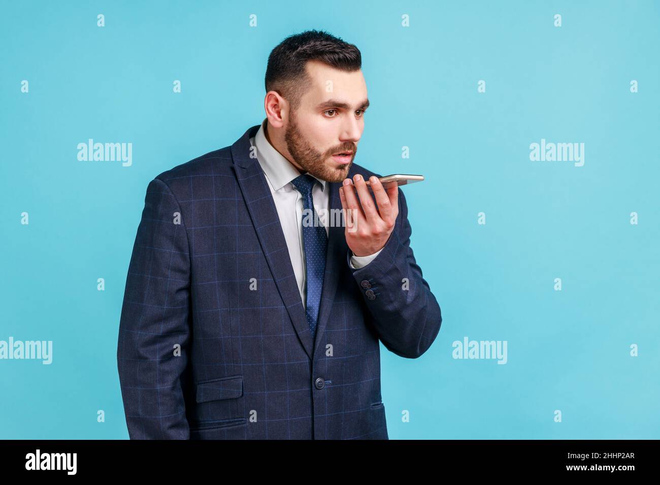 Uomo d'affari serio portatore di vestito di stile ufficiale che tiene il telefono cellulare in mano, dando comandi all'assistente vocale, lasciando il messaggio vocale. Studio interno girato isolato su sfondo blu. Foto Stock