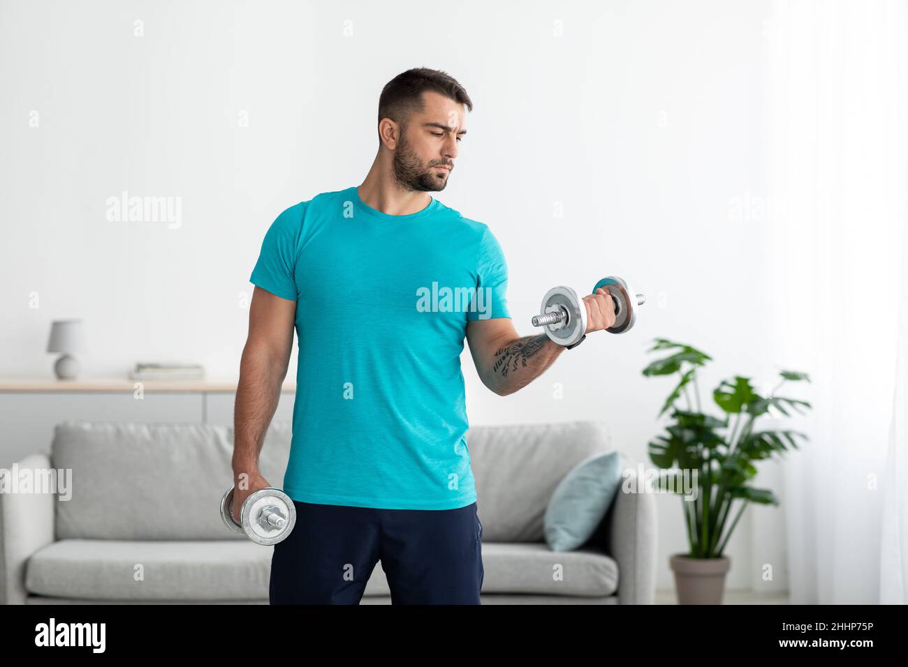 Concentrato giovane uomo caucasico atleta in t-shirt blu solleva manubri e fa esercizi per bicipite Foto Stock