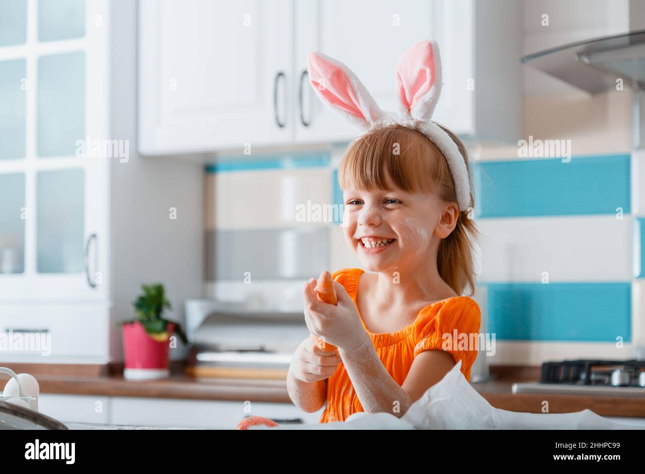 Ritratto emozionale di ragazza allegra vestita come coniglietto con carota per Pasqua mentre cucinano cibo in cucina a casa. Ragazza bambino che si diverte Foto Stock