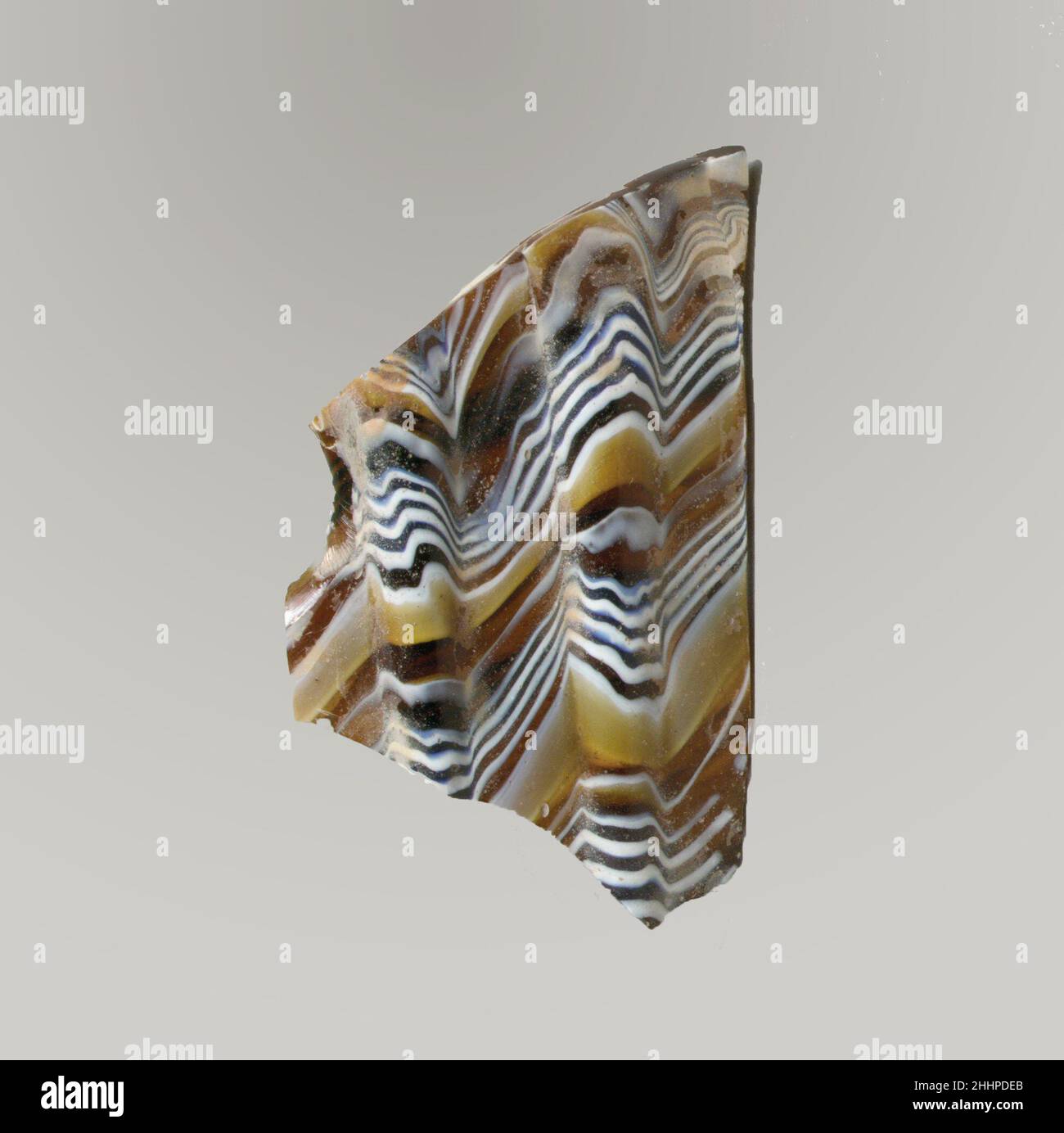 Mosaico di vetro dentato frammento di ciotola fine 1st secolo a.C.–inizio 1st secolo d.C. frammento di corpo romano.marrone di miele traslucido, blu di cobalto e bianco opaco.collo leggermente soprastato; Lato diritto rastremato verso il basso e curvato in basso. Motivo a mosaico a nastro formato da sezioni di una canna singola in marrone macinato con fili ondulati irregolari bianchi e blu in linee parallele; all'esterno, due ampie nervature verticali, abbastanza distanziate, con sommità appiattita e bordi esterni arrotondati.interno lucido; vaiolatura di bolle superficiali all'interno; sbucciature e macchie di cremoso iridescente agenti atmosferici all'esterno Foto Stock