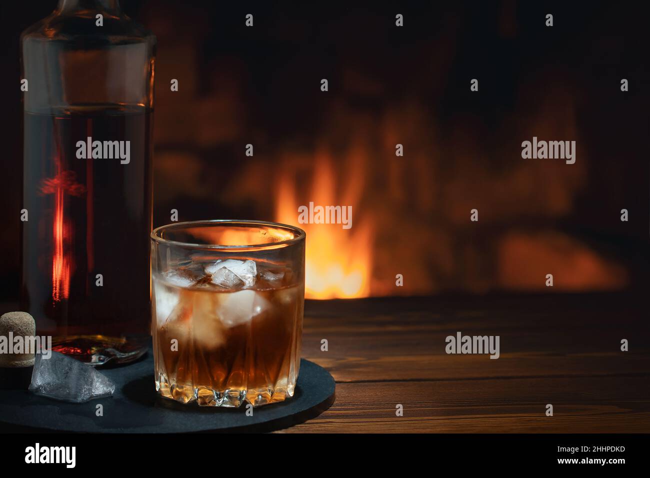 Bottiglia e bicchiere di whiskey con ghiaccio sul tavolo vicino al camino. Concetto di riposo e relax Foto Stock